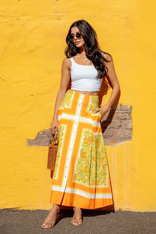 Woman in a white top and orange floral skirt standing against a yellow wall.