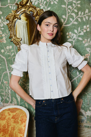 A woman modeling a white short-sleeve blouse with puffed sleeves and a high gathered neckline, paired with dark blue jeans, standing in front of a green floral wallpaper background with an ornate gold mirror.